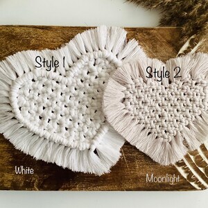 May include: Two heart-shaped macrame coasters with fringe edges. The left coaster is labeled Style 1 and White, featuring a bright white knotted cord pattern. The right coaster is labeled Style 2 and Moonlight, featuring a soft beige knotted cord pattern.