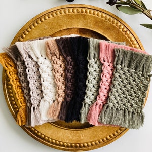 May include: A collection of rectangular macrame coasters arranged in a fan shape, featuring a variety of colors including mustard yellow, grey, white, beige, dark brown, black, sage green, and dusty pink, each with fringed ends and knotted patterns.