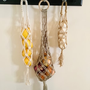 May include: Three macrame hanging produce bags with wooden rings. The bags are filled with lemons, onions, and garlic. The bags are hanging from hooks on a black shelf.