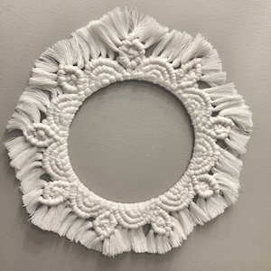 May include: White macrame wall hanging with a circular shape and a fringe border. The macrame is made with a knot pattern that resembles a flower.