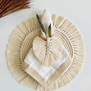 May include: A white cotton napkin with a beige border is placed on a round, beige macrame placemat with fringe. A wooden napkin ring with a knotted heart design holds the napkin in place. A sprig of dried lavender is tucked into the ring.