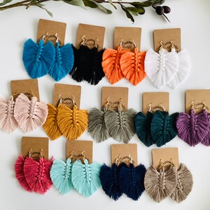 May include: Assortment of handmade tassel earrings in various colors, including teal, orange, white, pink, mustard yellow, gray, green, purple, red, light blue, navy blue, and beige. Each pair of earrings features a heart-shaped design with a fringe tassel.