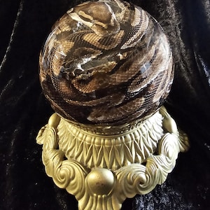 May include: A decorative crystal ball with a snake skin pattern, resting on a gold-colored ornate stand. The ball features a detailed print of brown and tan snake scales. The stand has a detailed, vintage design.