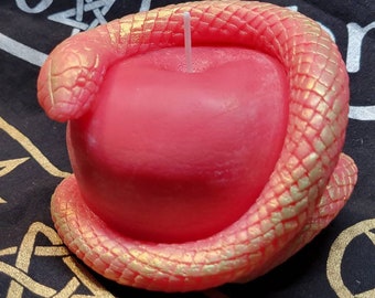 May include: A red apple-shaped candle wrapped in a coiled snake with textured scales. The snake features a gradient of pink and gold metallic tones. A single white wick protrudes from the center top of the red wax apple, creating a detailed sculpture.