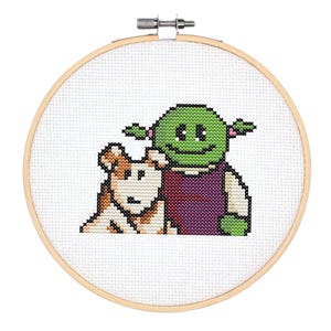 May include: Cross-stitch embroidery hoop with a green ogre character with pink hair and a brown dog. The ogre is wearing a purple dress and has a smiling face.