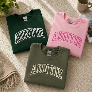 Three t-shirts with the word "Auntie" printed on them in different colors - green, pink, and olive - laid out on a carpeted floor.