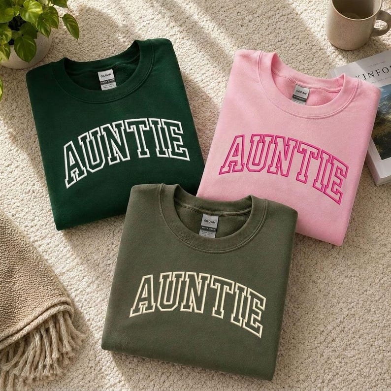 Three t-shirts with the word "Auntie" printed on them in different colors - green, pink, and olive - laid out on a carpeted floor.