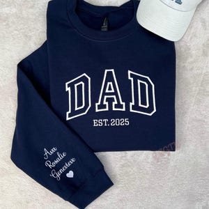 May include: Navy blue sweatshirt with the word "DAD" in white block letters, outlined in navy. The text "EST. 2025" is below the word "DAD". The left sleeve has the names "Ava Rosalie Genevieve" and a heart. A white baseball cap is in the image.