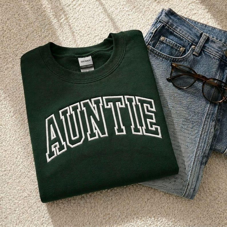 A green sweatshirt with the word "AUNTIE" printed on it, along with a pair of blue jeans and a pair of glasses.