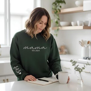 A woman in a green sweatshirt stands in a kitchen, holding a mug and looking down at a notebook on the counter.