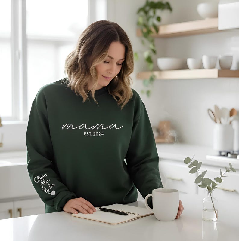 A woman in a green sweatshirt stands in a kitchen, holding a mug and looking down at a notebook on the counter.