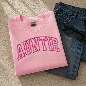 A pink sweatshirt with the word "AUNTIE" printed on it, along with a pair of blue jeans and a pair of glasses.