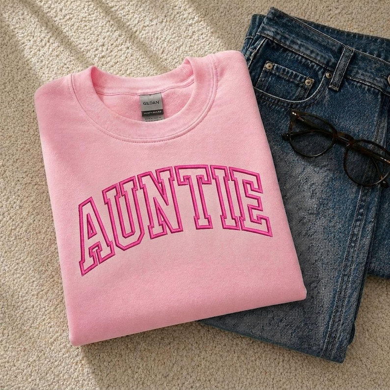 A pink sweatshirt with the word "AUNTIE" printed on it, along with a pair of blue jeans and a pair of glasses.