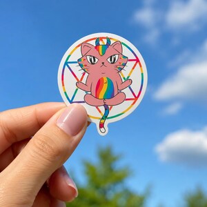 May include: A circular sticker featuring a pink cat with rainbow-colored horns, ears, and a belly patch. The cat is sitting in a meditative pose in front of a colorful geometric star design. The sticker has a white border and a glossy finish.