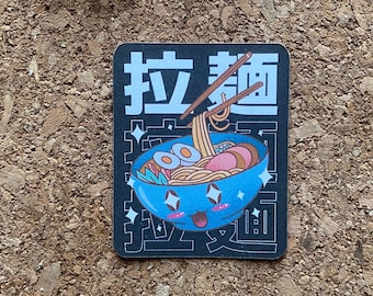 Op de afbeelding: Een zwart-witte sticker met een cartoonillustratie van een kom ramennoedels met eetstokjes. De tekst "拉麺" staat in Japanse karakters op de sticker.