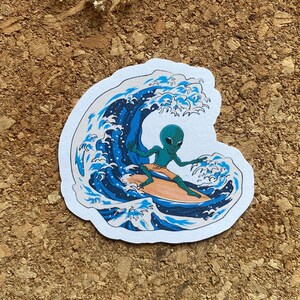 May include: A sticker featuring a green alien surfing on a wave. The wave is blue and white with a Japanese-inspired design.