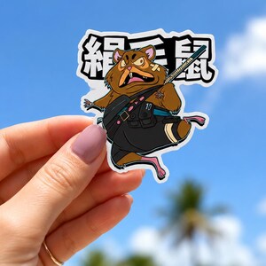 May include: A die-cut sticker featuring a brown cartoon hamster with an angry expression, wearing a black tactical vest with the number 10, and carrying a staff. Behind the character are large black Japanese characters on a white background.