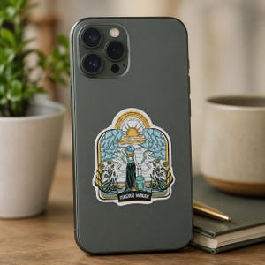 May include: A dark gray smartphone featuring a sticker with an illustration of a person standing with a suitcase, facing a sun, clouds, and flowers. The sticker includes the text "FOREVER WANDER" in a black banner at the bottom of the design.