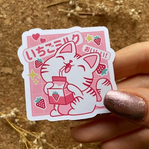 Pode incluir: Um adesivo rosa e branco com um gato de desenho animado bebendo leite de morango. O adesivo tem o texto japon&ecirc;s "いちごミルク" que se traduz como "leite de morango" e "おいしい!" que se traduz como "delicioso!"