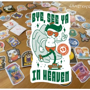 Op de afbeelding: Een sticker met een cartoon engel op een skateboard, met de tekst "BYE, SEE YA IN HEAVEN". De engel draagt een zonnebril en een rode zwembroek. Verschillende kleurrijke stickers omringen de hoofdsticker.
