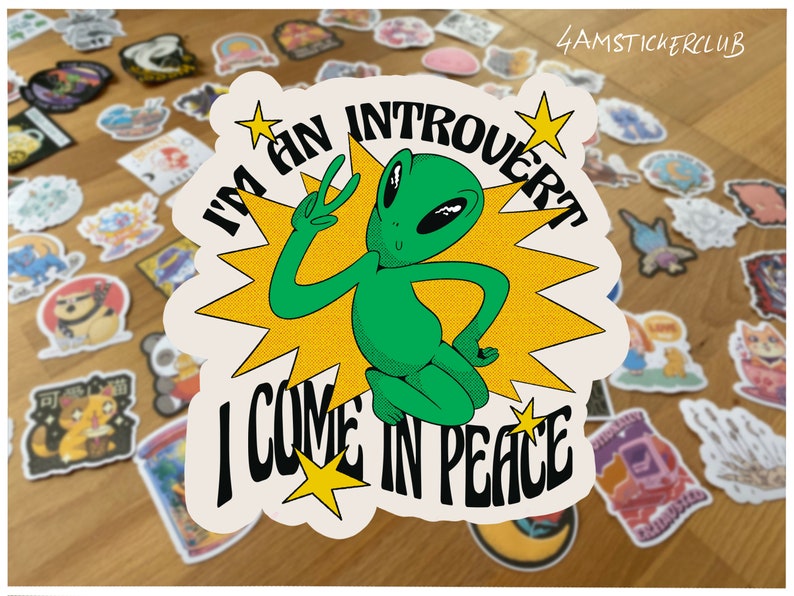 Pu&ograve; includere: Un adesivo bianco con un personaggio alieno verde che fa il segno della pace. L'adesivo ha stelle gialle e il testo "Sono un introverso, vengo in pace".