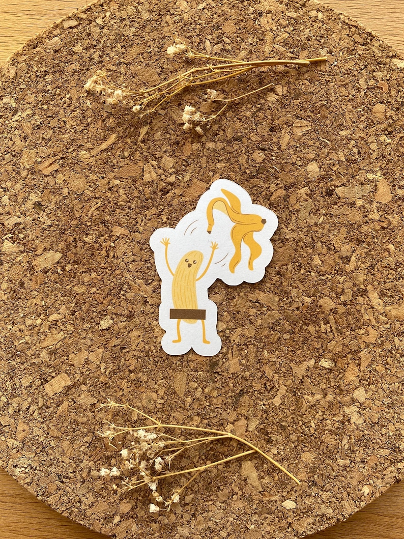 May include: A sticker of a banana with a face and arms, peeling itself. The banana is yellow and the peel is brown. The sticker is on a corkboard background.