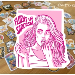 Pode incluir: Adesivo rosa com uma ilustração em estilo pop art de uma mulher com o texto "FLUENT IN SARCASM". O adesivo é rodeado por outros adesivos coloridos numa superfície de madeira.