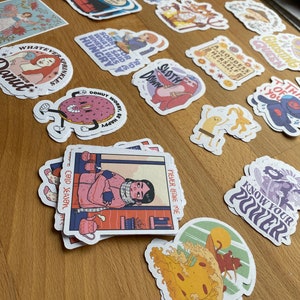 May include: A collection of colorful stickers featuring various illustrations and text. Some of the stickers include a donut with the text "Donut worry, be happy", a woman with the text "Whatever sprinkles", a sloth with the text "Sloth", and a person with the text "Cold Season".