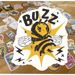Könnte beinhalten: Ein weißer Aufkleber mit einer Cartoon-Biene mit schwarz-gelben Streifen, einem wütenden Gesichtsausdruck und dem Wort "BUZZ" darüber. Die Biene ist von einem gelben Ausbruch mit Blitzen umgeben. Der Hintergrund ist mit verschiedenen bunten Aufklebern bedeckt.