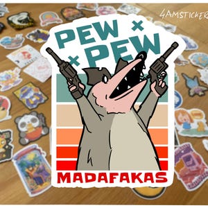 May include: A sticker with a cartoon opossum brandishing two guns, with the text "PEW PEW MADAF*KAS". The design uses a retro colour scheme with teal, beige, orange, and red stripes. Other stickers are visible in the background.