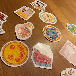 May include: Assortment of colorful stickers featuring various designs, including a pizza, a dragon, a skeleton hand holding a rose, a milk carton, a mermaid, a dinosaur, and a pig. The stickers are on a wooden surface.