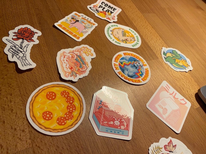 May include: Assortment of colorful stickers featuring various designs, including a pizza, a dragon, a skeleton hand holding a rose, a milk carton, a mermaid, a dinosaur, and a pig. The stickers are on a wooden surface.