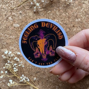 May include: A round sticker with a black background and orange text that reads "Feeling Devilish Don't Get Too Close". The sticker features a purple and white skull with horns and a cross behind it.