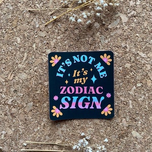 May include: Black sticker with white, pink, and blue text that reads "It's not me, It's my Zodiac Sign" with flower accents.