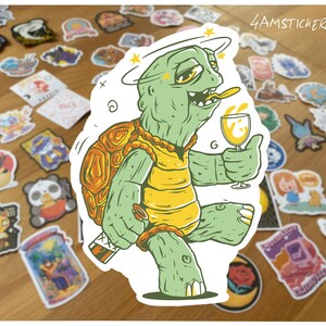 May include: A sticker depicting a cartoon turtle with a dizzy expression, holding a glass of wine and a small bottle. The turtle is green and yellow, with a gold shell and a halo. The sticker is surrounded by other stickers on a wooden surface.
