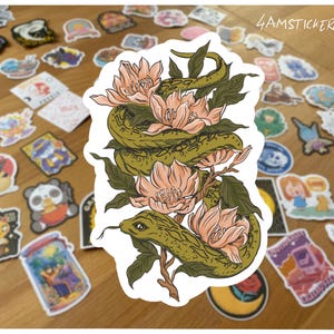 Könnte beinhalten: Ein Aufkleber mit einer grünen Schlange, die sich um pfirsichfarbene Blüten und grüne Blätter windet. Der Aufkleber trägt den Text "4AMSTICKERCLUB". Der Hintergrund ist eine Holzoberfläche mit verschiedenen Aufklebern.