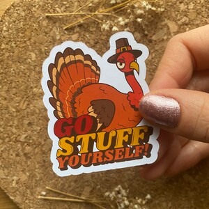 Op de afbeelding: Een witte sticker met een cartoon-kalkoen die een pelgrimhoed draagt. De kalkoen heeft een boze uitdrukking en zegt "Go Stuff Yourself!"