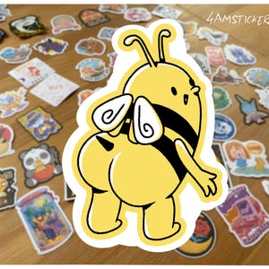 Può includere: Un adesivo a forma di ape dei cartoni animati giallo e nero con un contorno bianco. L'ape è rivolta verso l'esterno. L'adesivo è circondato da altri adesivi su una superficie di legno. Il testo "4AMSTICKERCLUB" si trova nell'angolo in alto a destra.