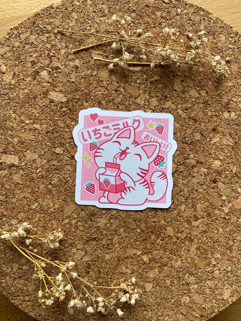 Pode incluir: Um adesivo rosa e branco com um gato de desenho animado bebendo leite de morango. O gato est&aacute; usando uma coleira rosa e tem uma express&atilde;o feliz. O adesivo diz "いちごミルク" em japon&ecirc;s, o que significa "leite de morango", e "おいしい!" que significa "delicioso!"