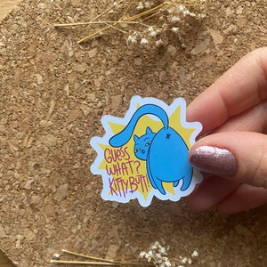 Può includere: Adesivo di un gatto blu con sfondo giallo a forma di stella. L'adesivo presenta il testo "Guess What? Kitty Butt!" in rosa e giallo. Il gatto è rivolto verso destra con la coda alzata.