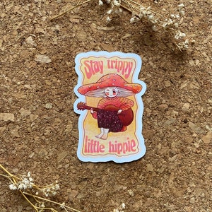 May include: A sticker with a cartoon illustration of a person with a mushroom cap, playing a guitar. The text "Stay trippy little hippie" is written in a retro font.