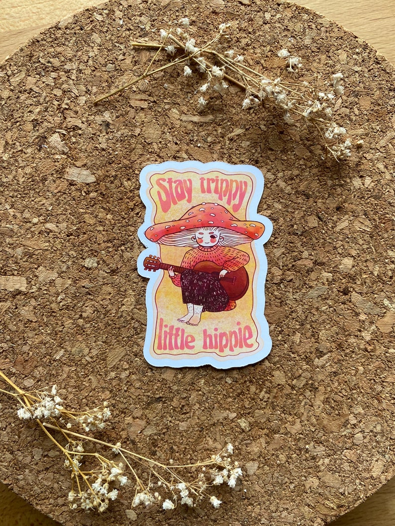 May include: A sticker with a cartoon illustration of a person with a mushroom cap, playing a guitar. The text "Stay trippy little hippie" is written in a retro font.