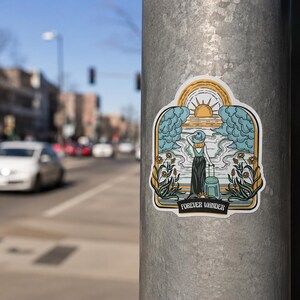 May include: A die-cut sticker featuring a stylized illustration of a person wearing a hat and holding a suitcase, standing before a sun, clouds, and flowers. The design includes a banner at the bottom with the text FOREVER WANDER in black.