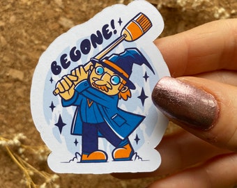 Op de afbeelding: Een witte sticker met een cartoonillustratie van een tovenaar die een bezem vasthoudt en een blauwe mantel en een punthoed draagt. De tovenaar vliegt door de lucht en de tekst "BEGONE!" staat boven hem.