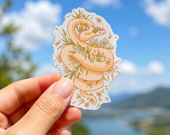 Può includere: Un adesivo fustellato raffigurante un serpente color pesca chiaro avvolto a spirale. Il serpente è decorato con piccole foglie verdi e fiori bianchi simili a margherite con centri gialli. L'illustrazione presenta un bordo bianco e una finitura opaca.