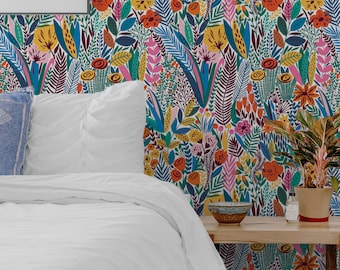 Op de afbeelding: Een wit bed met een wit dekbedovertrek en een blauw kussen. Het bed staat voor een muur met een kleurrijk bloemenbehang. Een klein houten bijzettafeltje met een plant en een kom erop staat naast het bed.