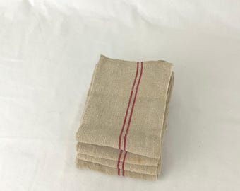 May include: A stack of four beige linen towels with two vertical red stripes. The towels are folded and stacked neatly, showcasing a rustic, farmhouse aesthetic. The towels are made of a natural, textured fabric.