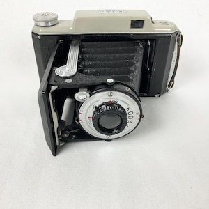 Puede incluir: Una cámara plegable Kodak vintage con cuerpo de cuero negro y lente plateada. La cámara tiene un dial blanco y negro con la palabra "Kodak" impresa en él.