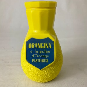 Può includere: Una bottiglia di plastica gialla a forma di arancia con un'etichetta blu che recita "Orangina à la pulpe d'Orange Pasteurisé".