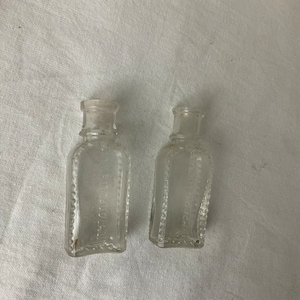 May include: Two small, clear glass bottles with a rectangular shape and rounded edges. The bottles have a beaded border and a narrow neck. The text on the bottles is partially visible. The bottles are on a white surface.